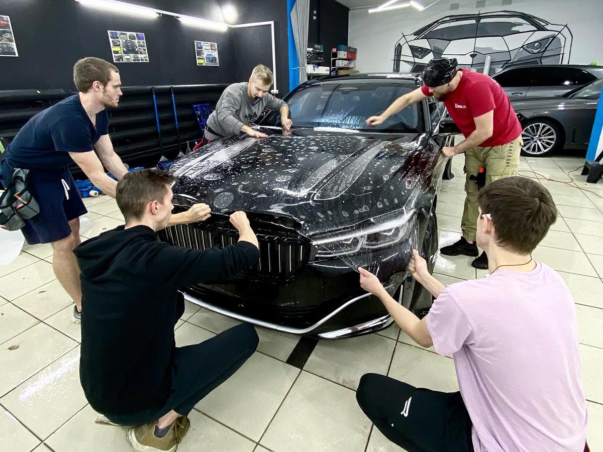 Смотреть на фото процесс оклейки антигравийной пленкой капота у машины BMW 7-ой серии.